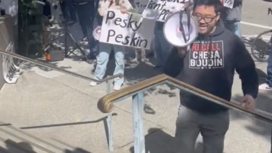A person holds a megaphone while others stand with signs in a protest on a city sidewalk.